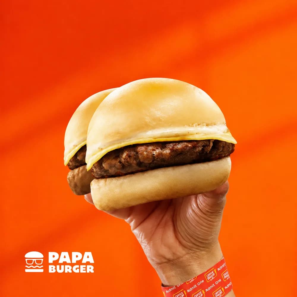 A hand holding two Papa Cheesy Burgers stacked together — buy one, take one.
